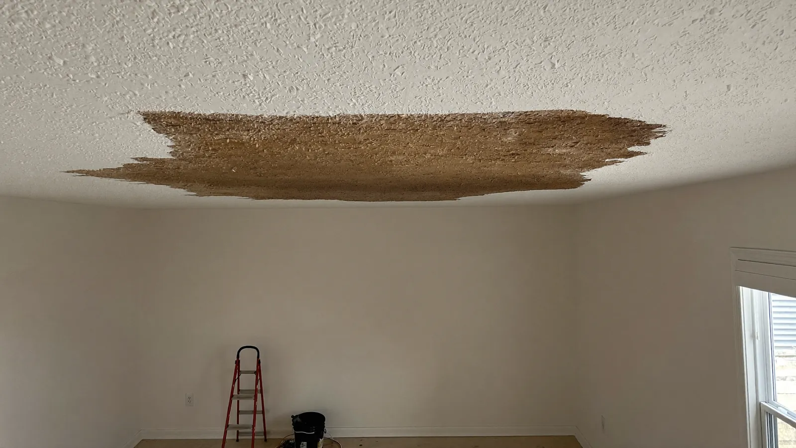Brown water stain spreading across a white interior ceiling below a roof leak, with a bucket on the floor underneath
