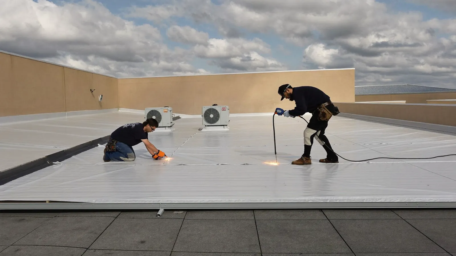 Worker heat-welding a seam on a white TPO membrane flat roof of a low commercial building, rooftop units and parapet in the background